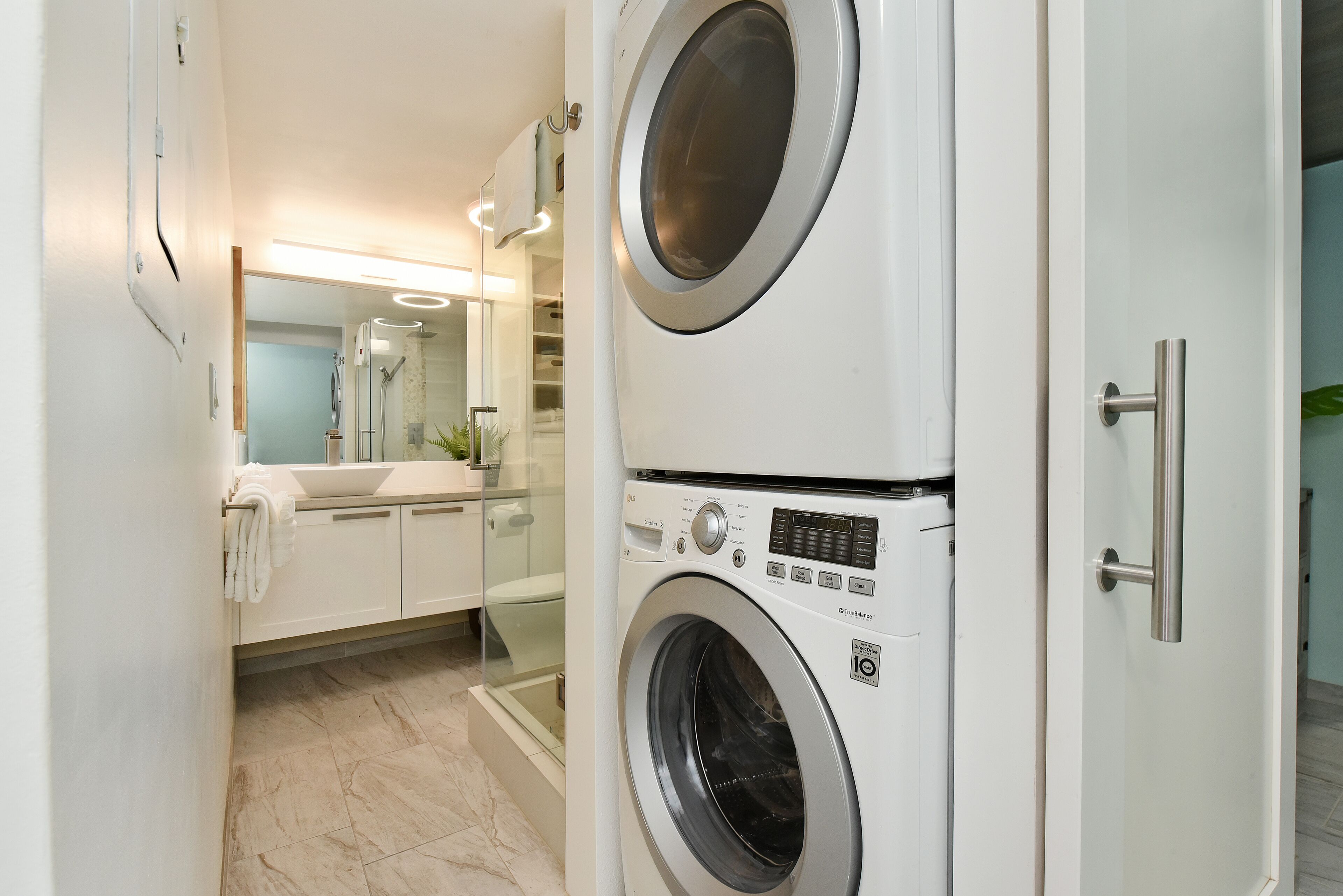 A modern laundry room with stacked washers and dryers, a glass shower enclosure, and a visible bathroom with a sink and towel rack.