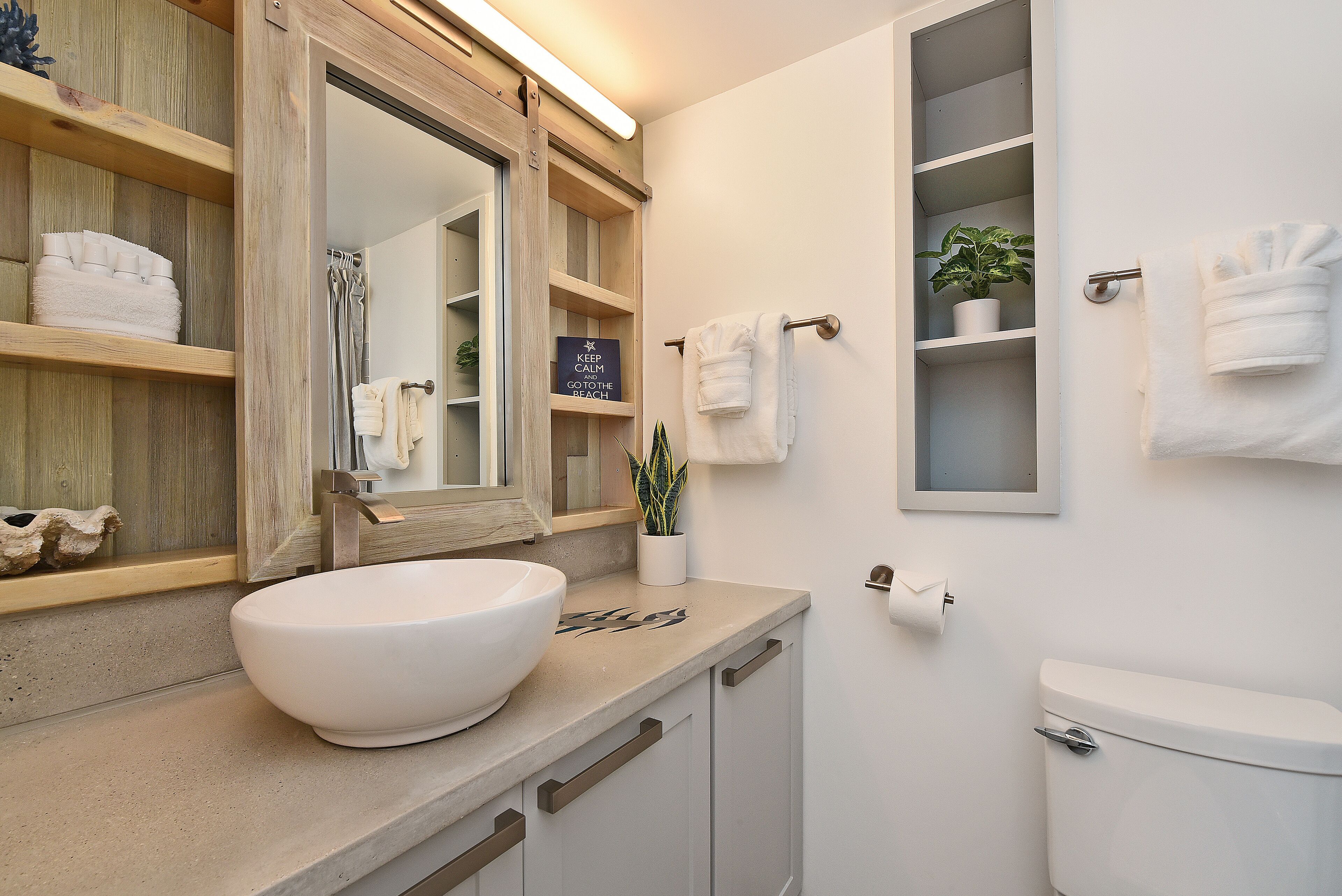 Ein Badezimmer mit einem weißen Waschbecken, Holzregalen, einem Spiegel, einem Handtuchhalter und einer Toilette.