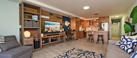 A modern living room with a grey sofa, a patterned rug, a TV, wooden shelving, and a kitchen area with stools.
