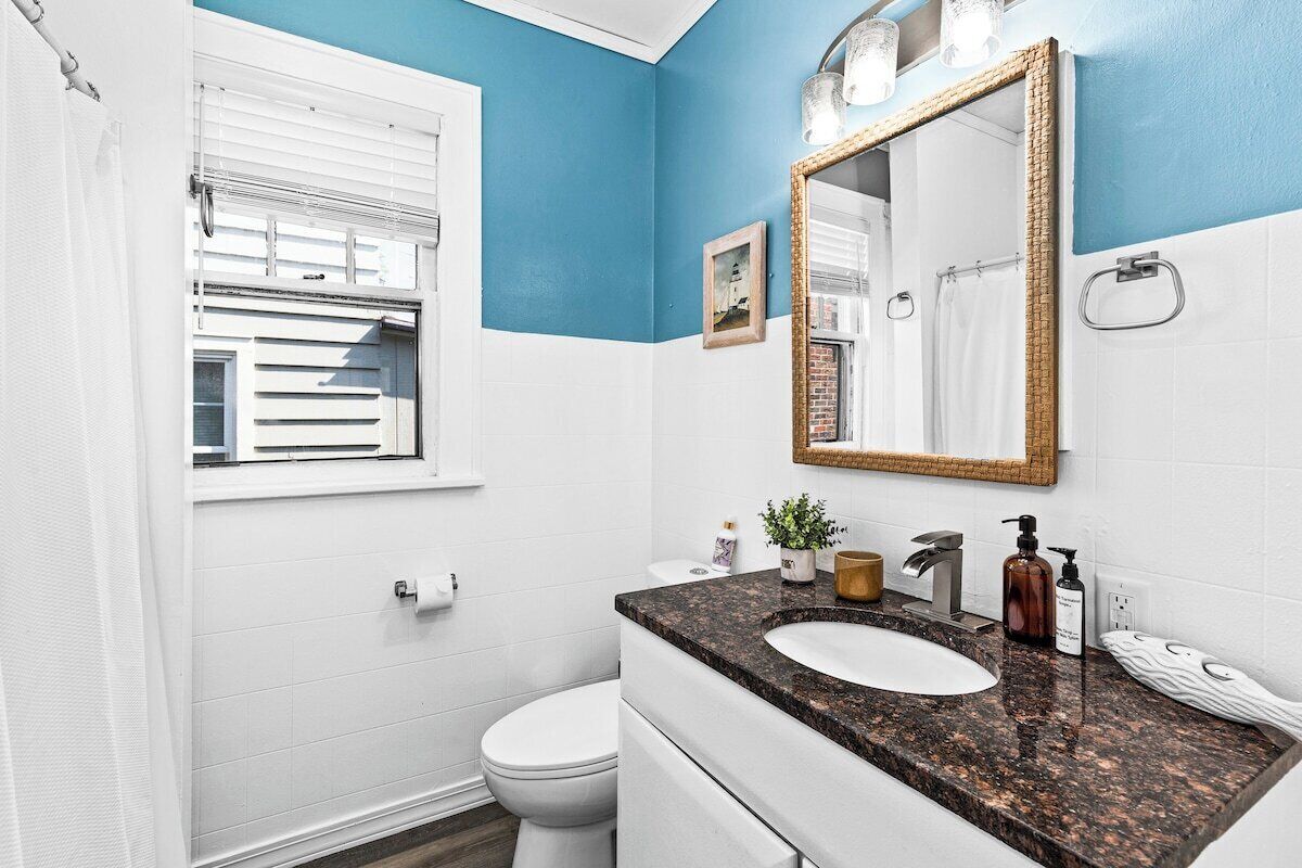 Un baño con inodoro blanco, lavamanos con encimera de granito, espejo con marco dorado y ventana con persianas.