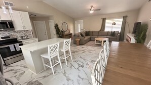 Eine moderne Küche mit eingebauter Mikrowelle und Backofen, eine Bar mit weißen Stühlen, ein Wohnzimmer mit Sofa und Couchtisch sowie ein Deckenventilator.
