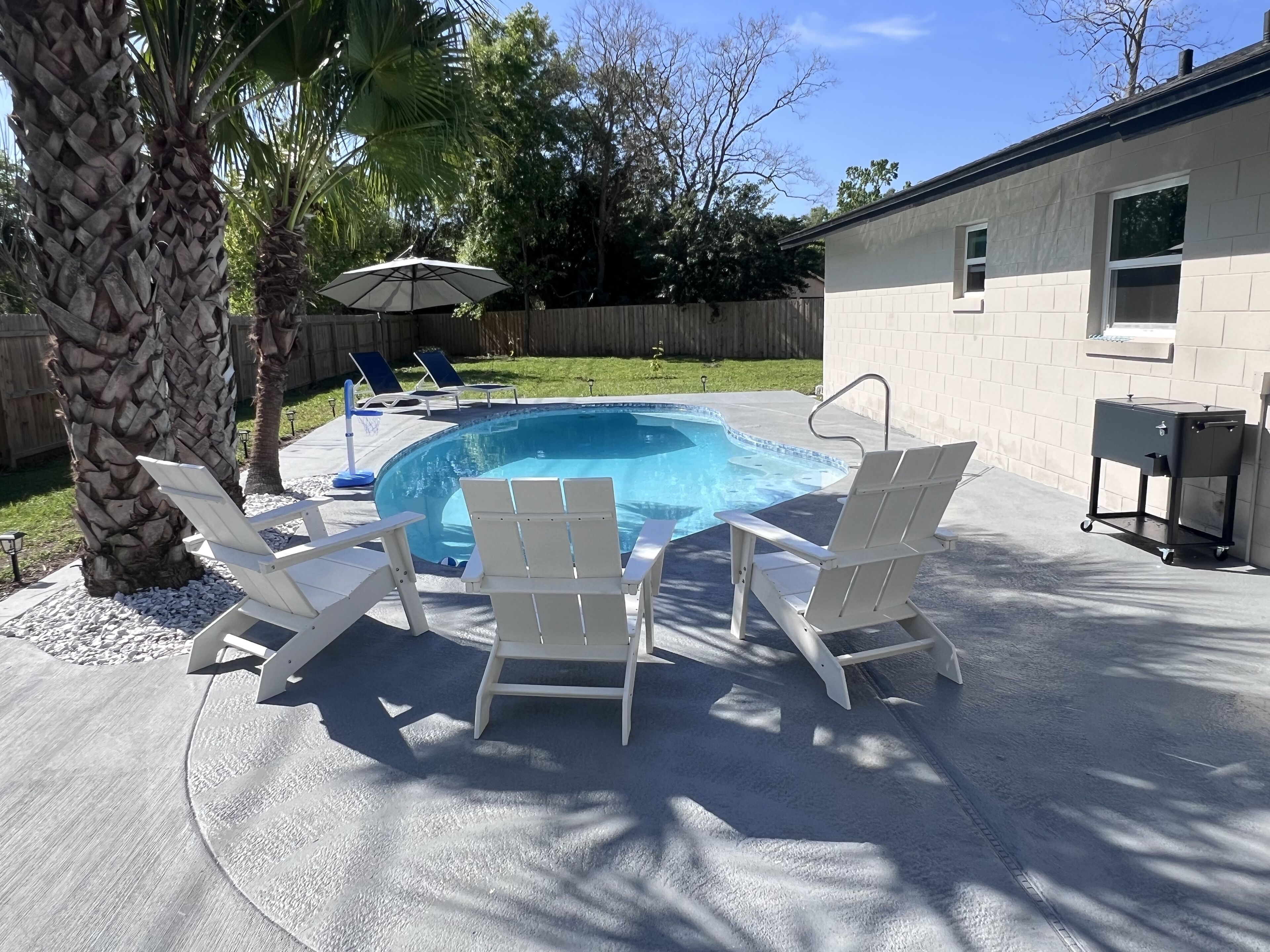 En trädgård med en bubbelpool, fyra vita solstolar, ett trästaket och ett hus.