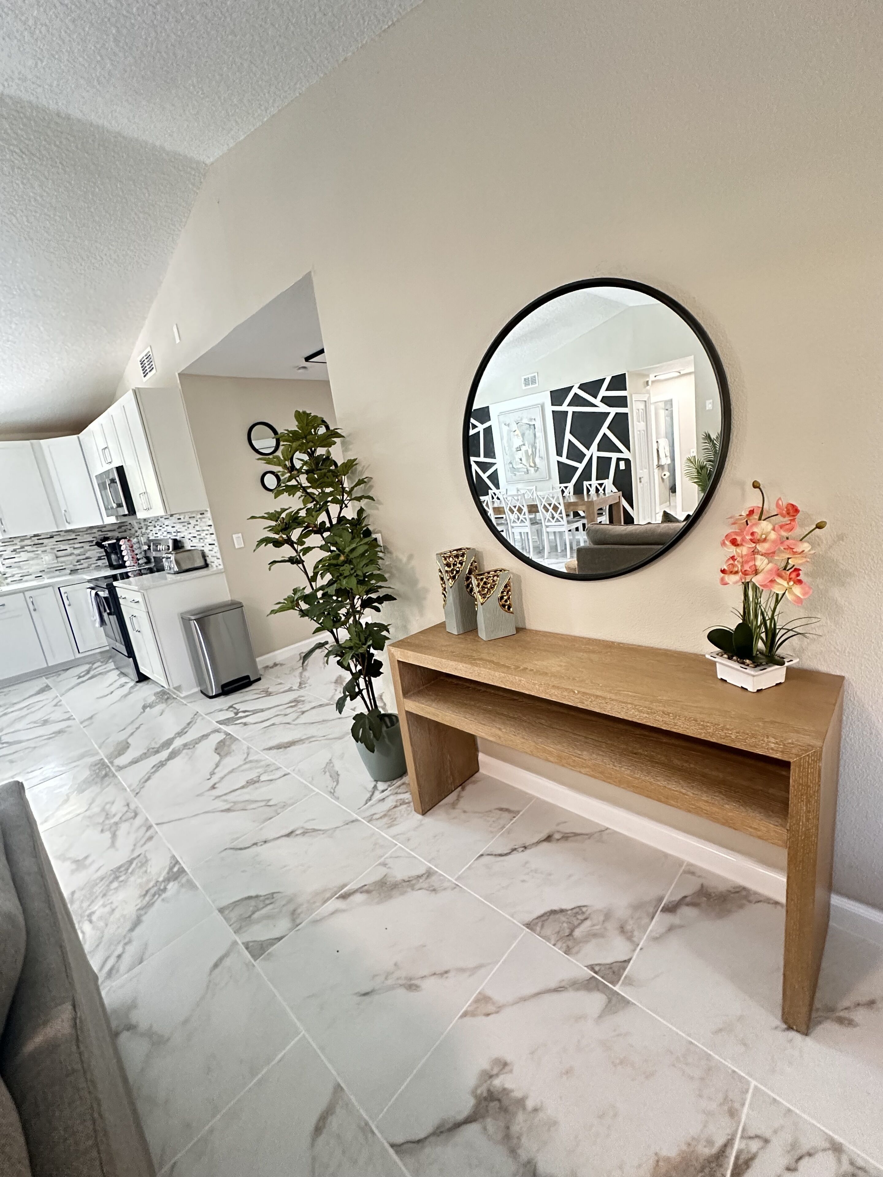 A modern kitchen with white cabinets, a marble floor, a round mirror, and a wooden bench with decorative items.