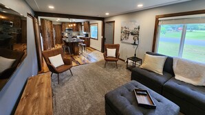 A spacious living area with a large window, a cozy seating arrangement, and a kitchen in the background. - Gorgeous 5 bed Remodel near Downtown Hudson for Groups! (Hudson)