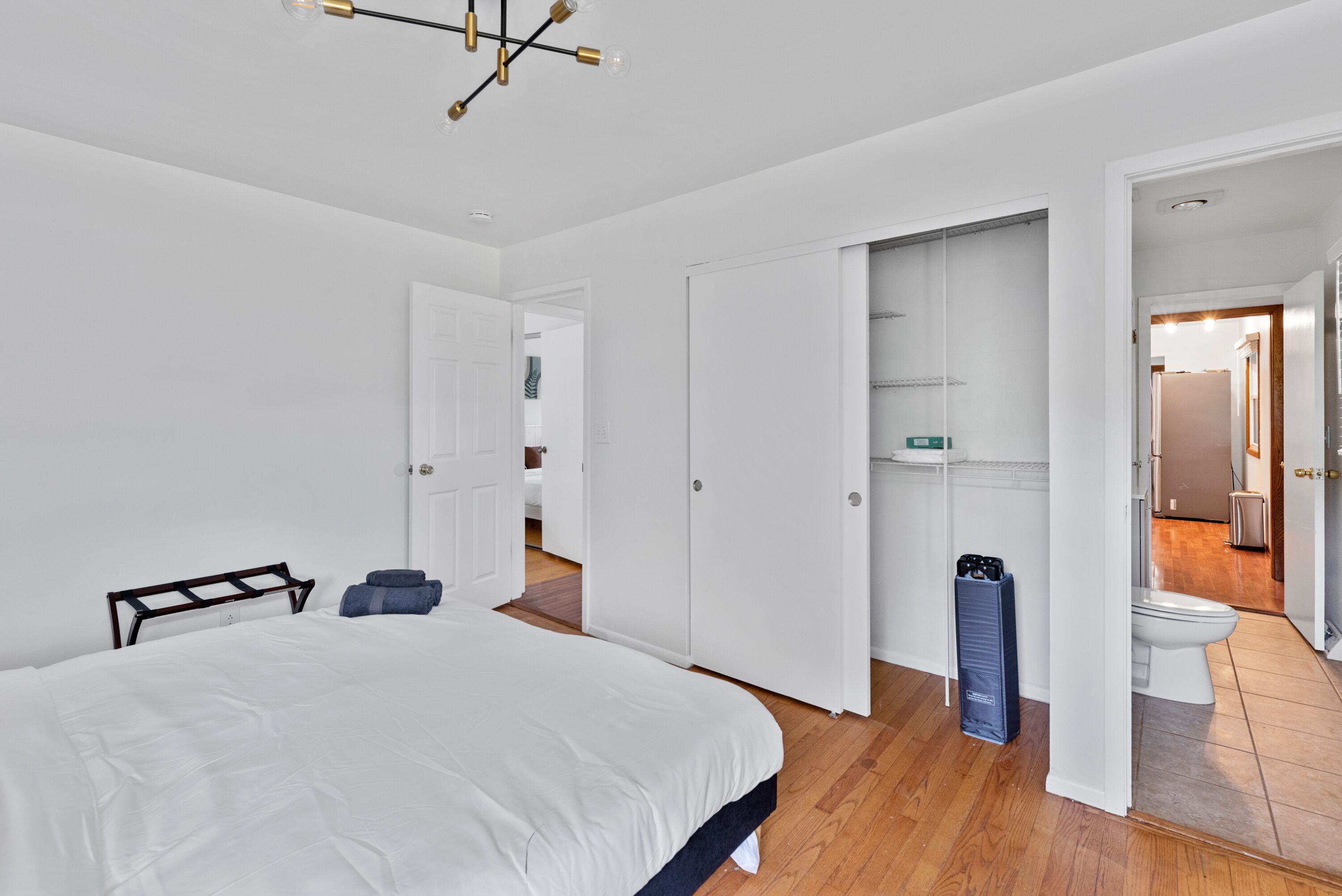 A modern bedroom with a large bed, white walls, and a wooden floor. There is a closet with open doors, revealing shelves and a hanging rack. A small heater is visible on the floor.