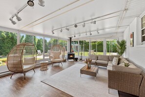 Uma sala de estar ampla com lareira, móveis de vime e janelas grandes com vista para o exterior.