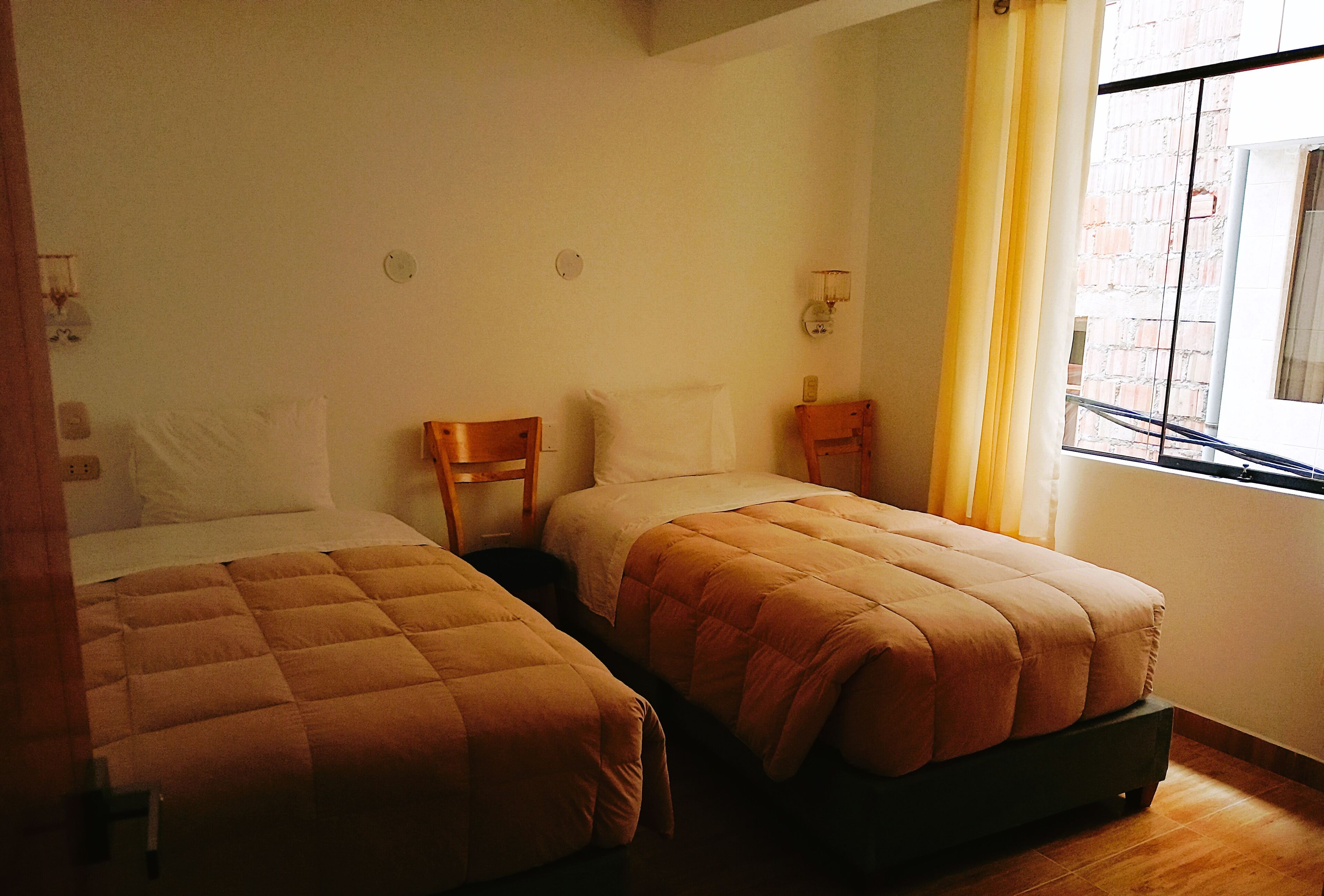 Una habitación con dos camas individuales, una silla de madera, una mesita y una ventana con vista a un edificio de ladrillo.