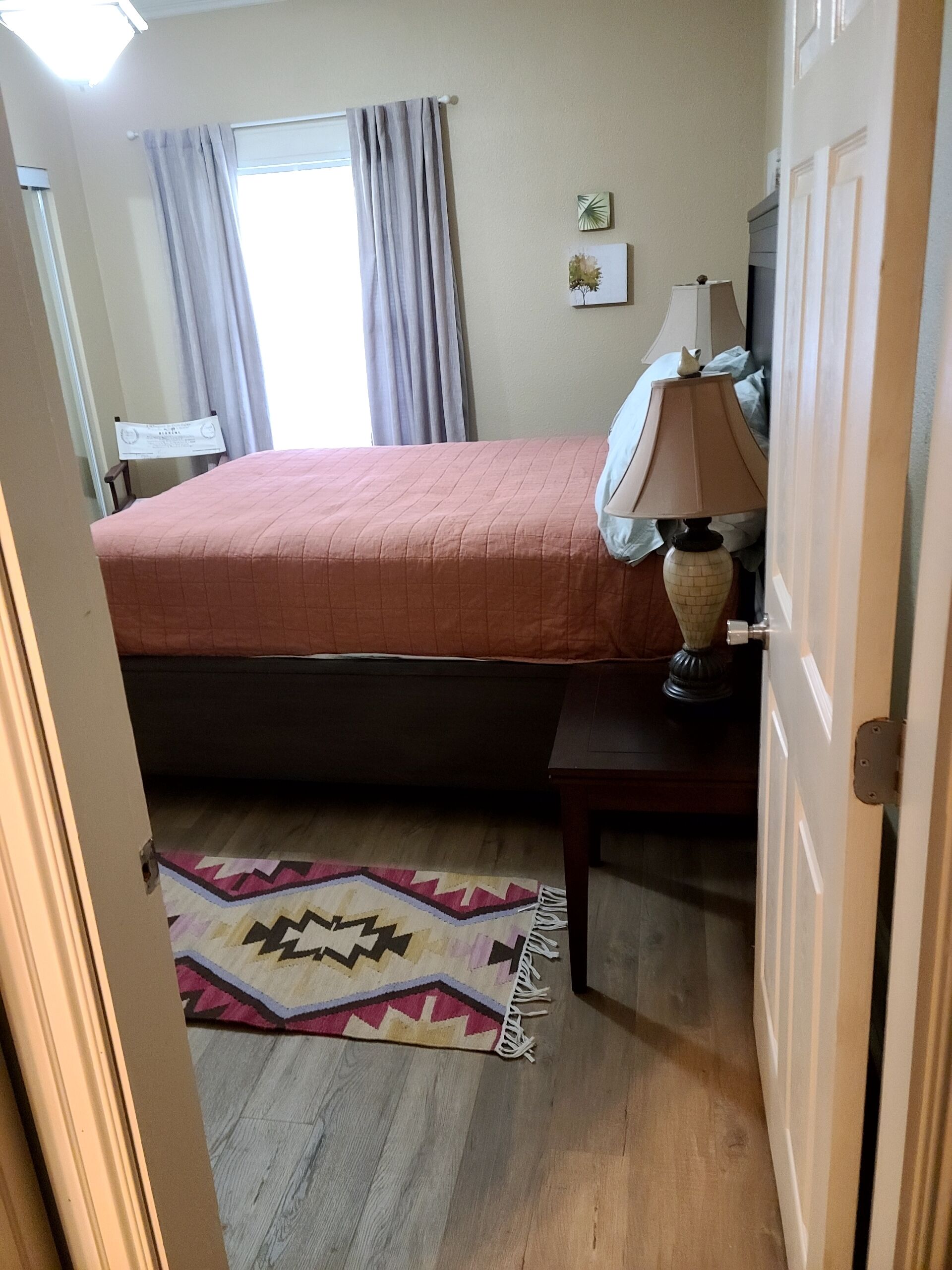A bedroom with a bed, a bedside table with a lamp, a rug, and a window with curtains.