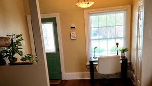 A living room with a wooden floor, a patterned rug, a green door, a white chair, and a small table with a book.