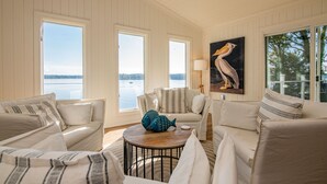 A spacious living room with a large window overlooking a body of water, a painting of a pelican, and a round coffee table with decorative fish.