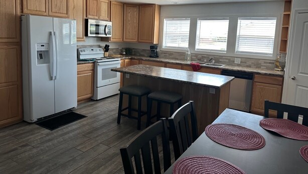 A modern kitchen with wooden cabinets, a white refrigerator, a microwave, and a stove. There is a window with blinds, a sink, and a dining area with a table and chairs. - For your best vacation ever! (Crescent City)