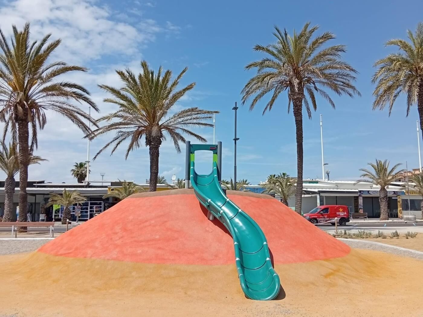 Un parque infantil con un tobogán verde, palmeras y un vehículo rojo al fondo.