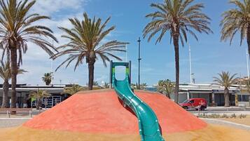 Un parque infantil con un tobogán verde, palmeras y un vehículo rojo al fondo.