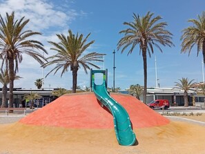 Un parque infantil con un tobogán verde, palmeras y un vehículo rojo al fondo.