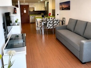 Ein modernes Wohnzimmer mit einem grauen Sofa, einem Essbereich mit einem gemusterten Tisch und einer Küche im Hintergrund.