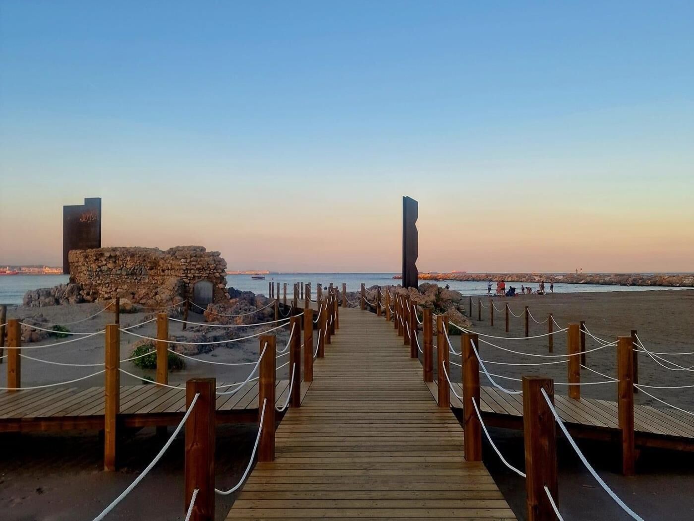 Una pasarela de madera que lleva a una playa con una estructura de piedra y un alto pilar a la derecha.