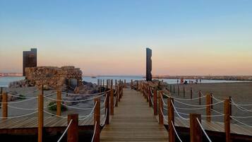 Una pasarela de madera que lleva a una playa con una estructura de piedra y un alto pilar a la derecha.