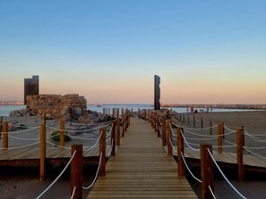 Una pasarela de madera que lleva a una playa con una estructura de piedra y un alto pilar a la derecha.