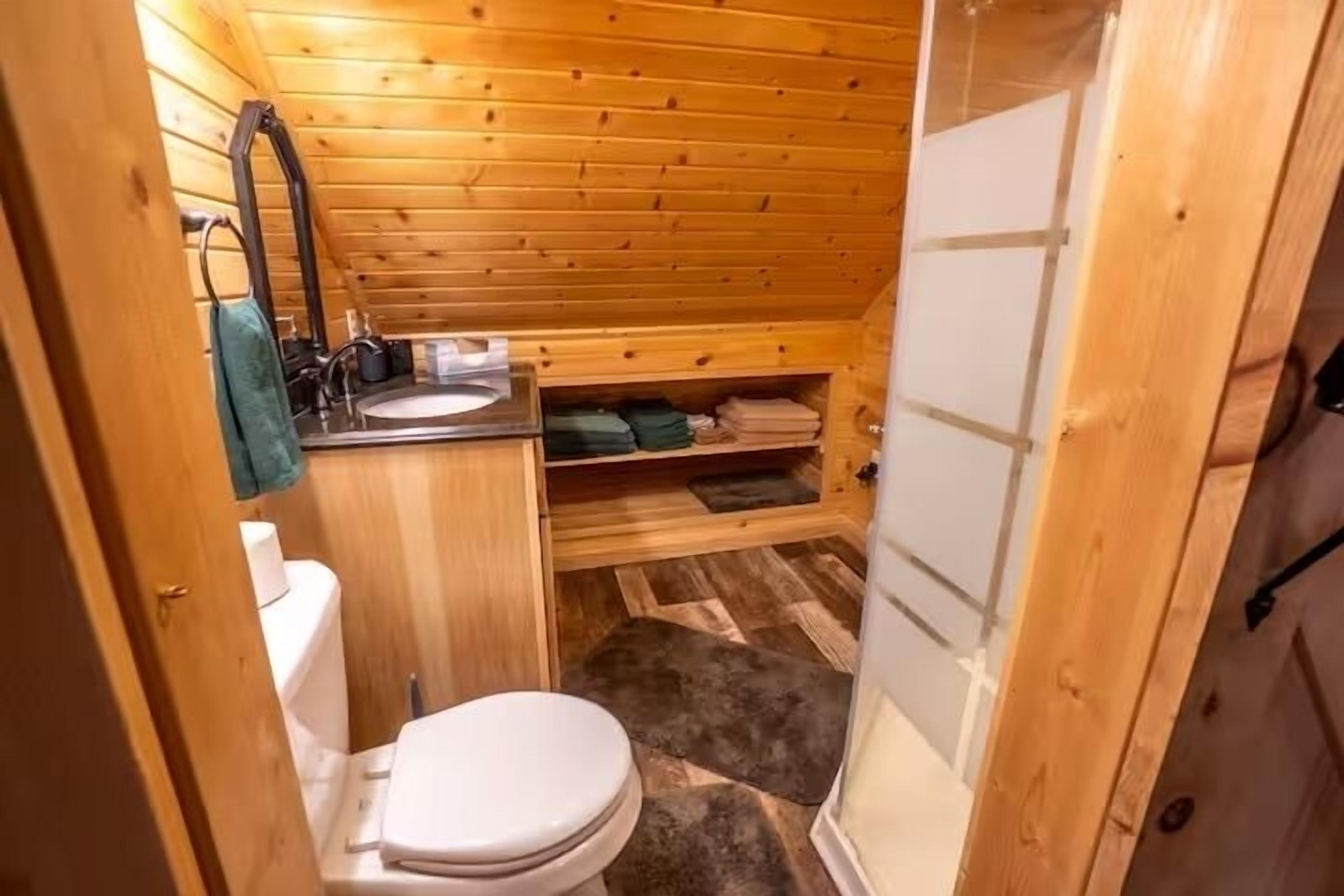 Baño de una cabaña de madera pequeña, con inodoro, lavamanos y un estante con toallas dobladas.