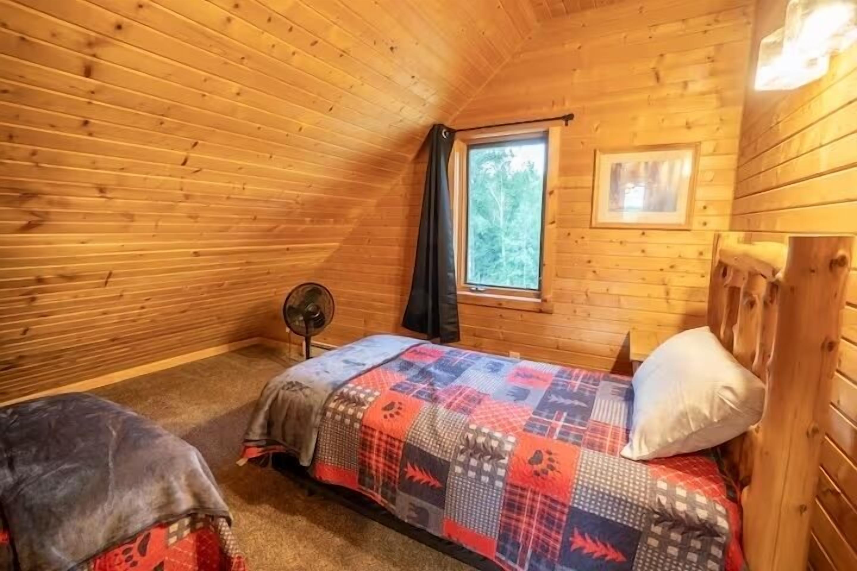 Ein gemütliches Blockhauskabinenzimmer mit Holzeinrichtung, einem Einzelbett mit gemustertem Bettbezug, einem kleinen Tisch und einem Fenster mit Blick auf Bäume.