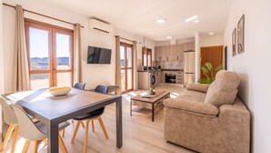 A modern living room with a sofa, a dining table with chairs, a kitchen area with cabinets and appliances, and a TV mounted on the wall.