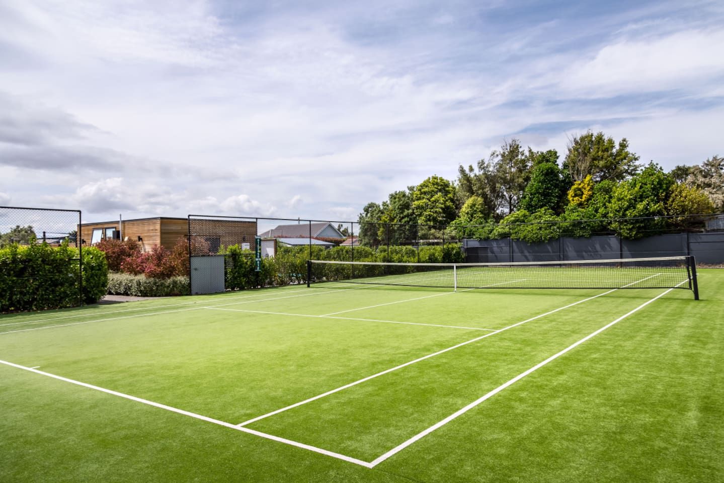 A tennis court with a net, surrounded by a fence and greenery.