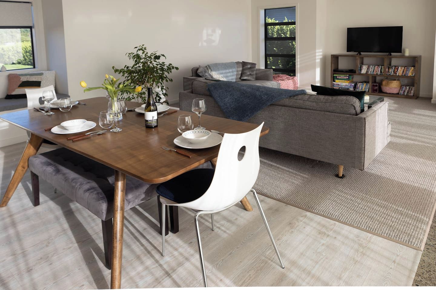 A modern living room with a dining table set for two, a grey sofa, a bookshelf, and a television.