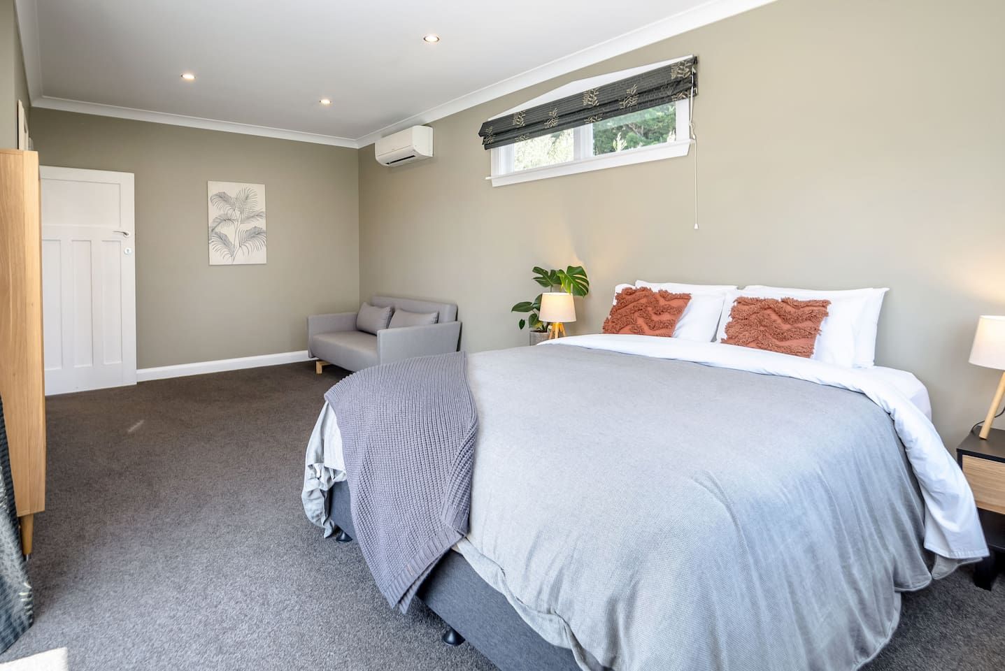 A bedroom with a large bed, a grey sofa, a window with blinds, and a wall-mounted air conditioner.