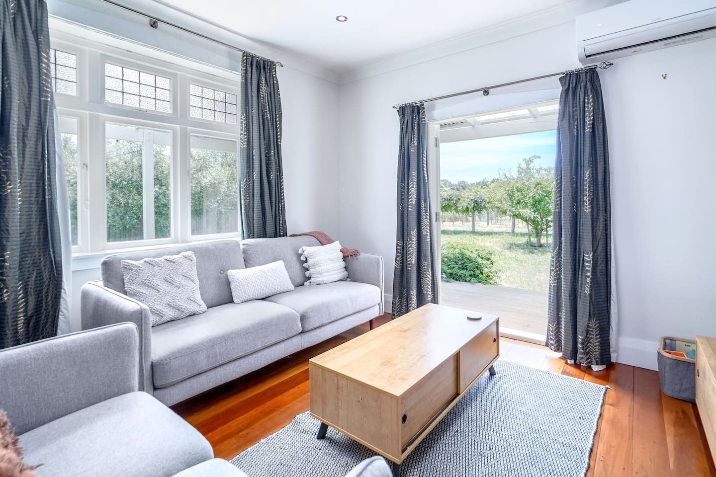A modern living room with a grey sofa, a wooden coffee table, and a view of greenery outside.