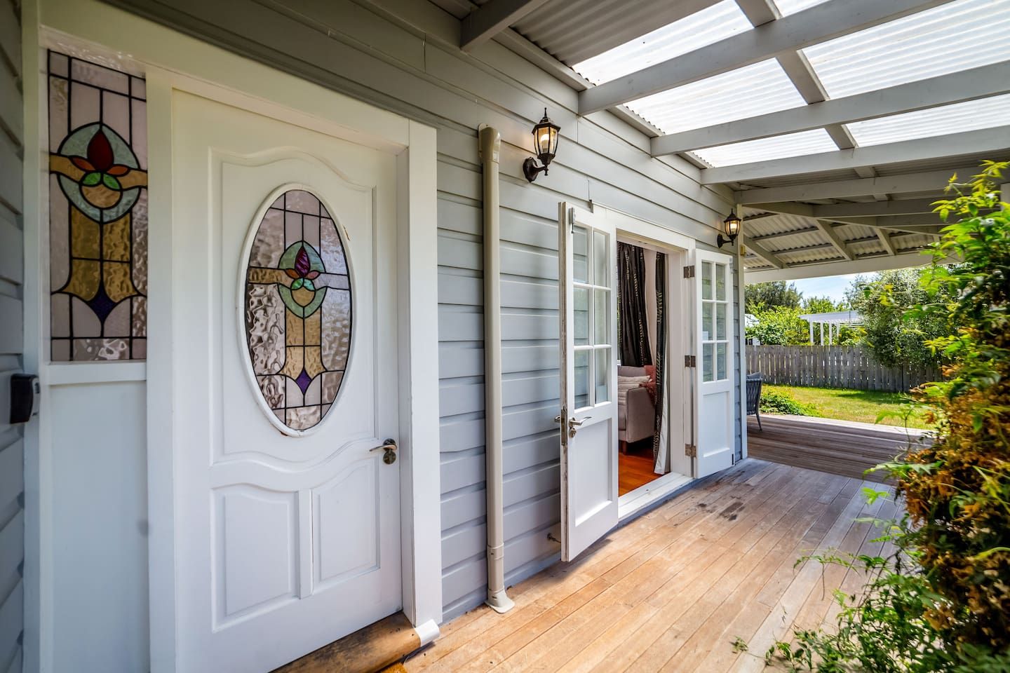 A covered porch with a wooden floor, a white door with stained glass, and a view of a backyard.