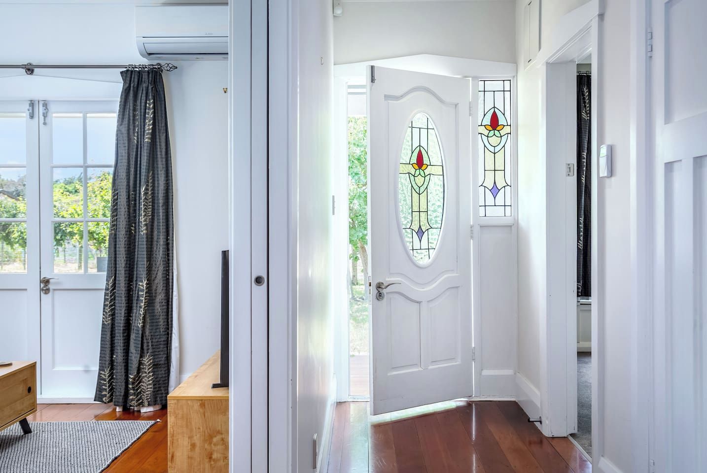 A hallway with a white door featuring stained glass windows, a wooden floor, and a view of greenery outside.