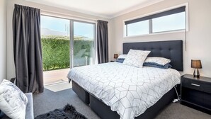 A bedroom with a large bed, a nightstand, a lamp, and a view of greenery outside. - 27 Tuscan, Martinborough (Martinborough)