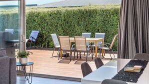 A modern outdoor dining area with a table and chairs, a potted plant, and a view of a green hedge and a building in the background. - 27 Tuscan, Martinborough (Martinborough)