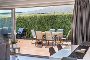 Un espace de restauration extérieur moderne, avec une table et des chaises, une plante en pot, et une vue sur une haie verdoyante et un bâtiment en arrière-plan.