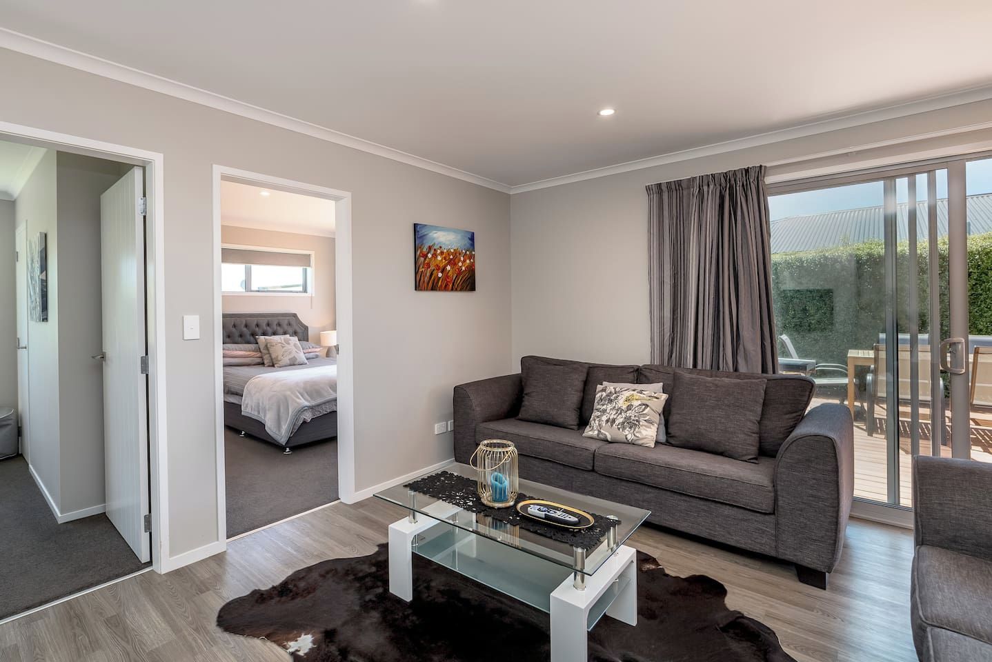 A modern living room with a glass coffee table, grey sofa, and a view of a balcony.
