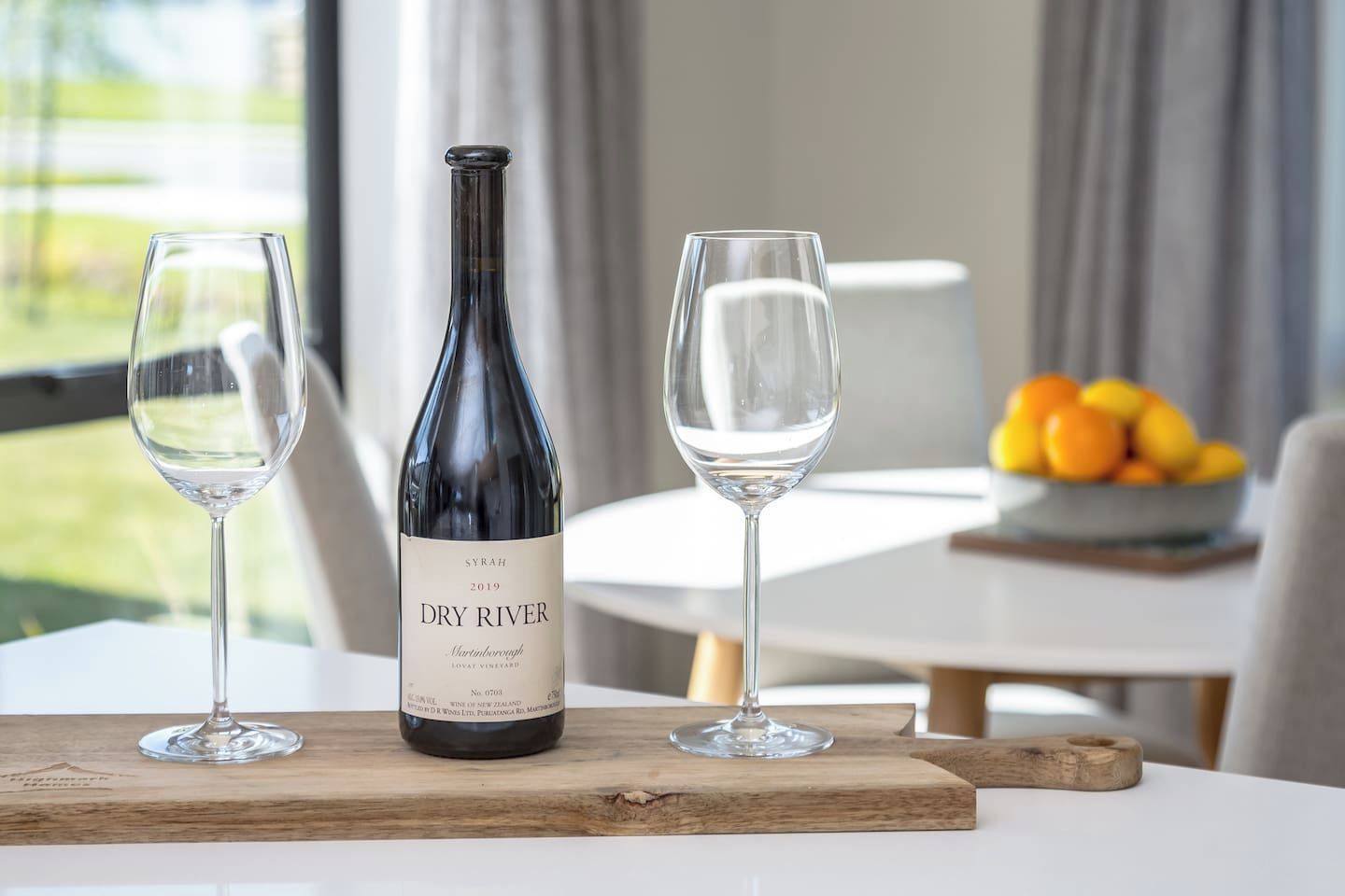 Una bottiglia di vino e due bicchieri su un tagliere, con una ciotola di frutta e un tavolo sullo sfondo.