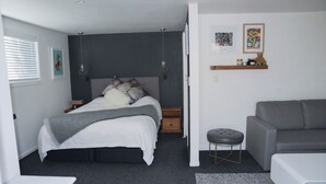 A bedroom with a bed, bedside tables, a grey sofa, a small stool, and artwork on the wall.