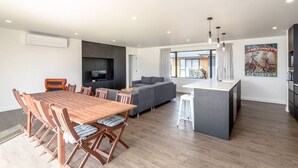 A modern living space with a wooden dining table, grey sofa, and a kitchen island. - 53 Burgundy – Large Group Retreat with Seasonal Communal Pool, Tennis & Expansive Deck (Martinborough)