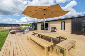 A wooden deck with picnic tables and a large umbrella, a barbecue grill, and a modern building with large windows.