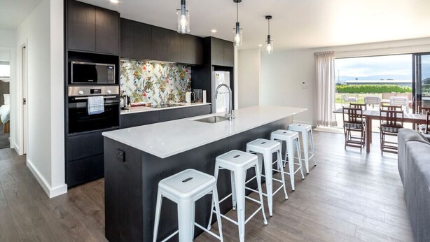 A modern kitchen with a central island, bar stools, and a dining area with a view. - 53 Burgundy – Large Group Retreat with Seasonal Communal Pool, Tennis & Expansive Deck (Martinborough)
