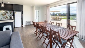 A modern kitchen with a white refrigerator, wooden dining table, and chairs. There is a large window with a view of the outdoors. - 53 Burgundy – Large Group Retreat with Seasonal Communal Pool, Tennis & Expansive Deck (Martinborough)