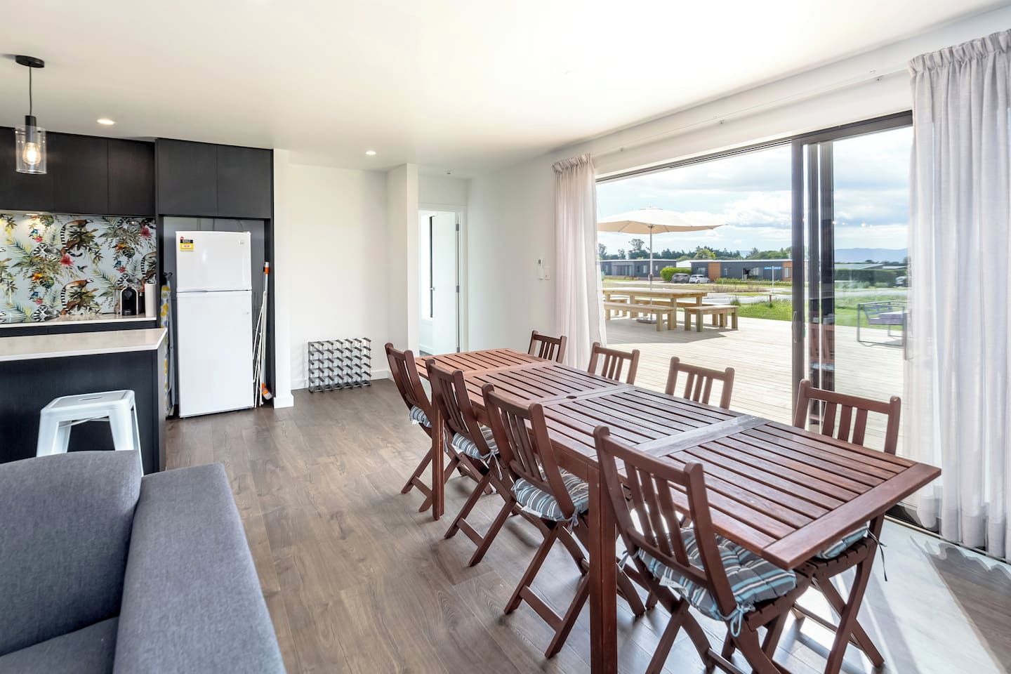 A modern kitchen with a refrigerator, a dining area with a wooden table and chairs, and a view of the outdoors through large windows.