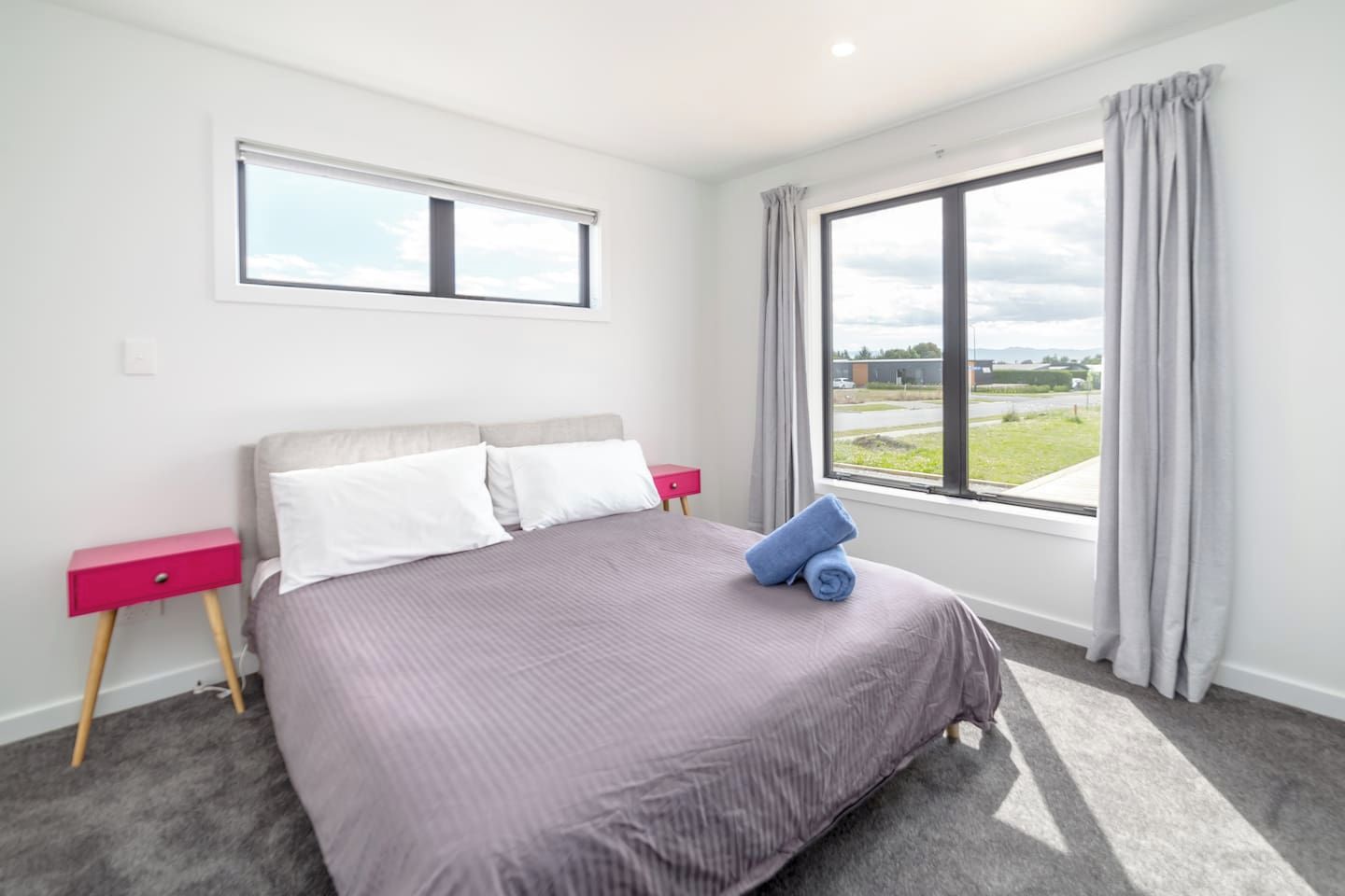 A bedroom with a bed, a nightstand, a window with curtains, and a view of the outdoors.