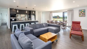 A modern living room with a grey sectional sofa, a wooden coffee table, and a red armchair. A kitchen with dark cabinets and a dining area with a table and chairs is visible in the background. - 53 Burgundy – Large Group Retreat with Seasonal Communal Pool, Tennis & Expansive Deck (Martinborough)