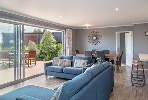 A modern living room with a blue sofa, dining table, and chairs, a large window with a view of greenery, and a decorative wall piece.