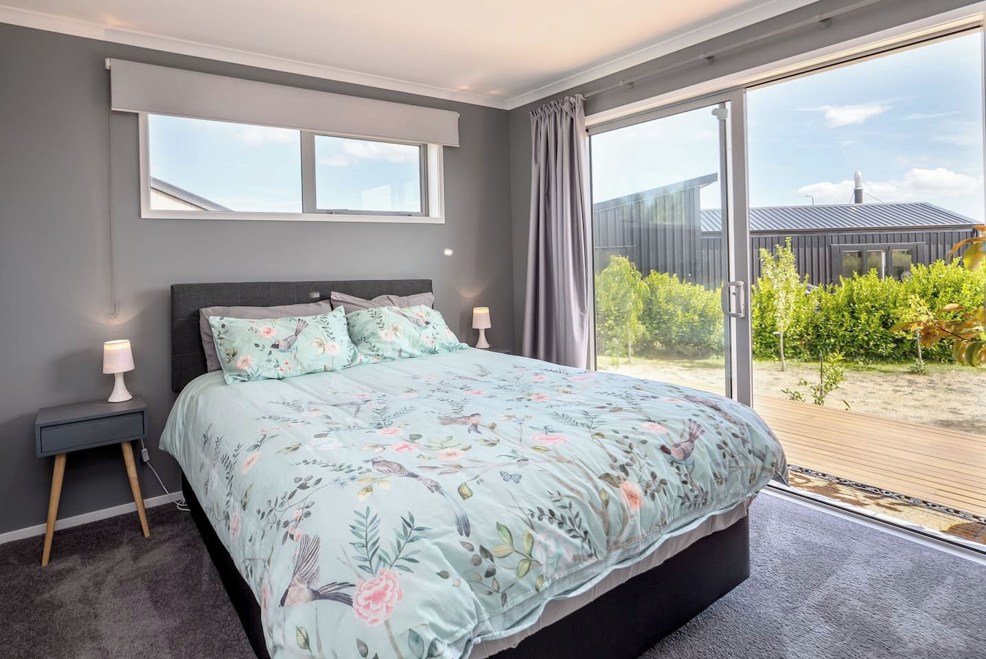 Ein Schlafzimmer mit einem großen Bett, einem Nachttisch, einer Lampe und Blick auf einen Garten durch eine Glastür.