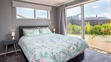 Ein Schlafzimmer mit einem großen Bett, einem Nachttisch, einer Lampe und Blick auf einen Garten durch eine Glastür.
