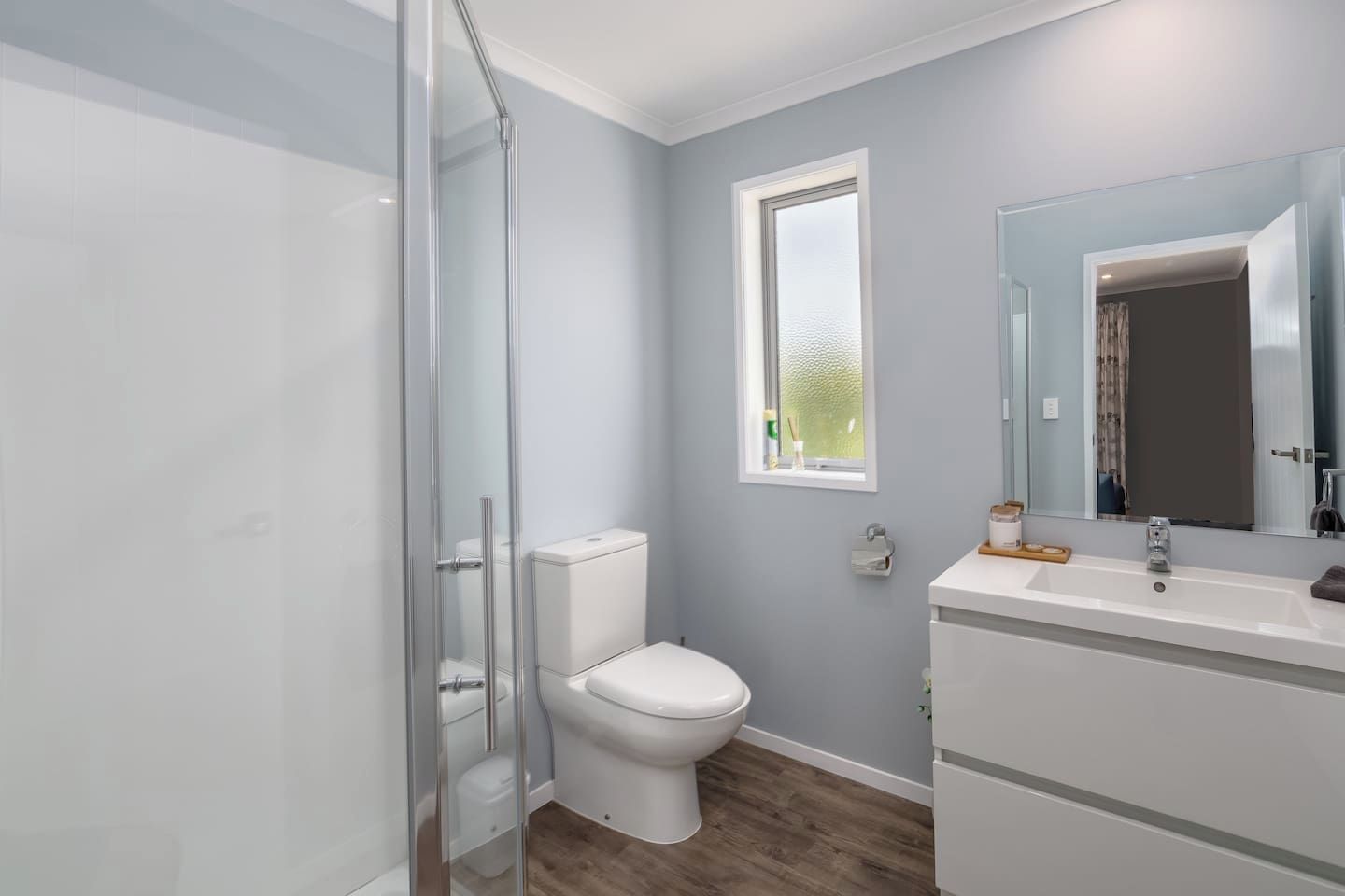 A bathroom with a glass shower enclosure, a white toilet, a window, and a sink with a mirror.