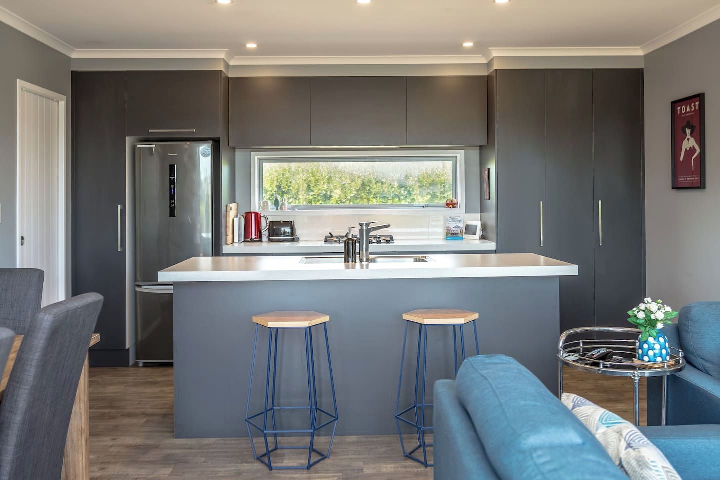 A modern kitchen with a central island, bar stools, and a view of greenery outside.