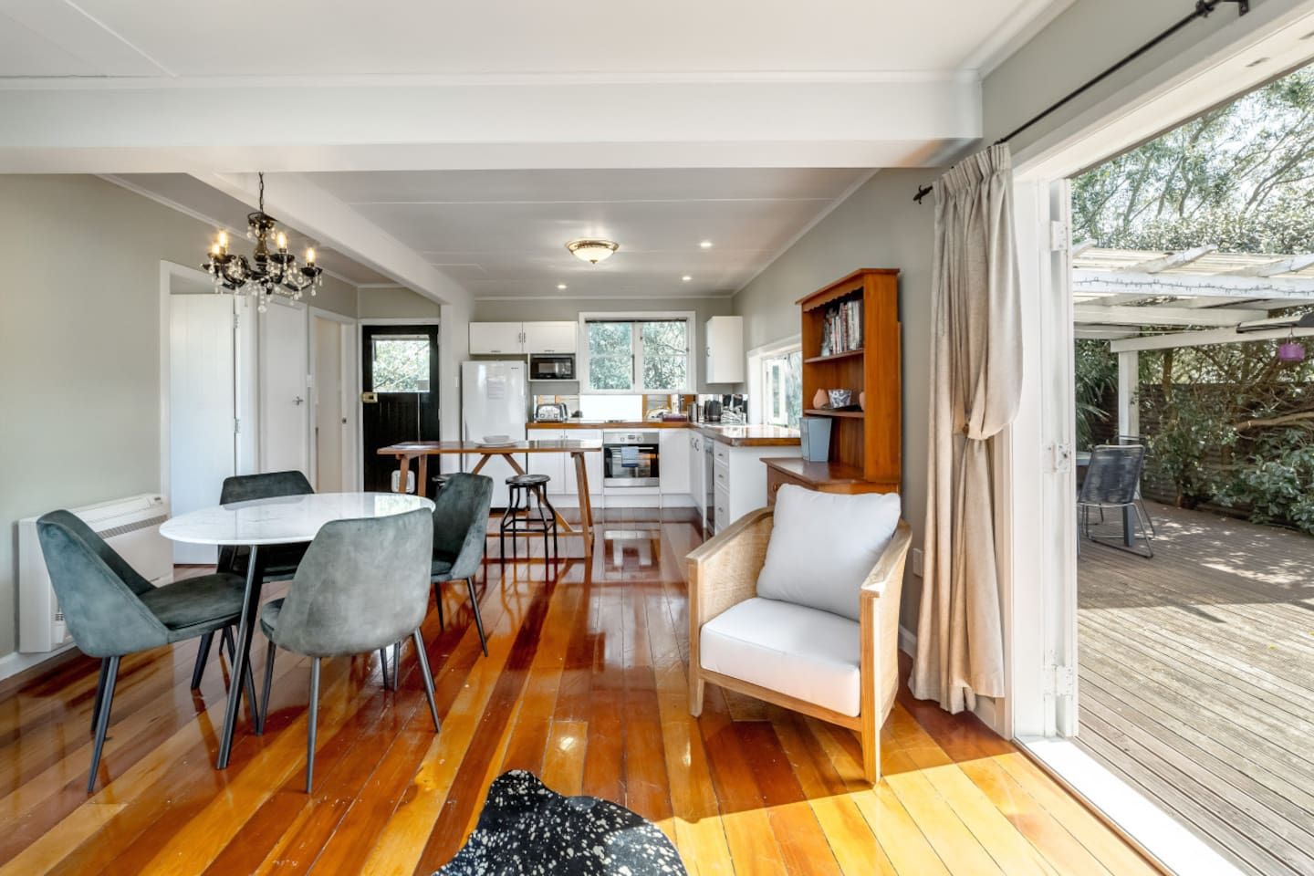 Una cocina moderna con armarios blancos, una zona de comedor con una mesa redonda y sillas, y una terraza de madera visible a través de un ventanal grande.